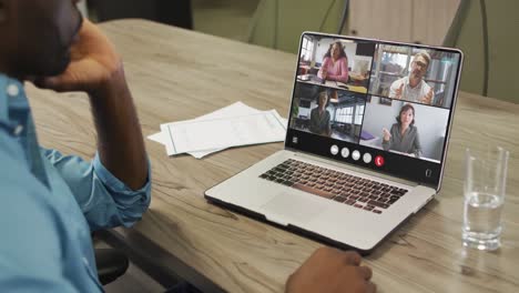 hombre afroamericano usando una computadora portátil para una llamada de video, con diversos colegas de negocios en la pantalla