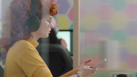 close-up-beautiful-young-redhead-business-woman-writing-notes-using-computer-planning-creative-project-texting-on-smartphone-in-colorful-modern-office-listening-to-music-wearing-headphones