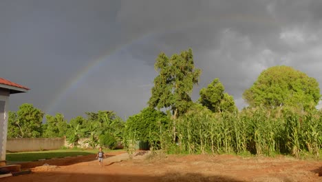 A-young-boy-runs-in-his-backyard-while-a-full-rainbow-is-in-the-sky-among-dark-storm-clouds-and-the-orange-glow-of-a-sunset-shines-on-the-trees