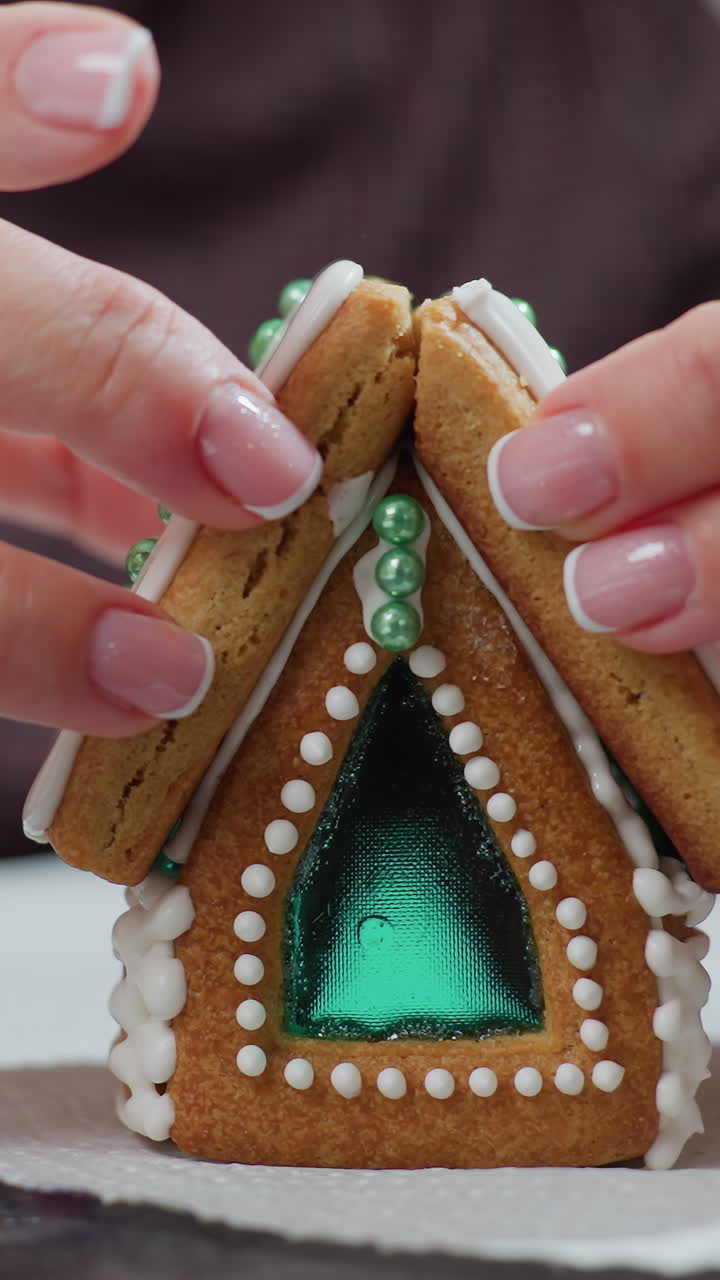 신선한 아이싱과 복잡한 장식으로 새로 조립된 진저브레드 하우스 쿠키를 안정시키는 베이커의 클로즈업, 하 킨에 제대로 설정되도록 구조를 조심스럽게 유지합니다.