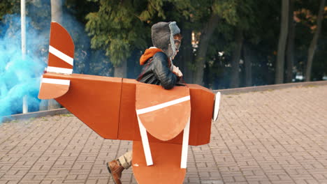 side view of a cute little running in the park holding a cardboard plane with smoke behind and playing as an aviator