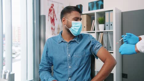 Portrait-Of-Handsome-Young-Hindu-Male-Patient-In-Medical-Mask-Sitting-In-Hospital-Room-Receiving-Coronavirus-Vaccine-In-Quarantine,-Senior-Male-Professional-Doctor-Doing-Vaccination