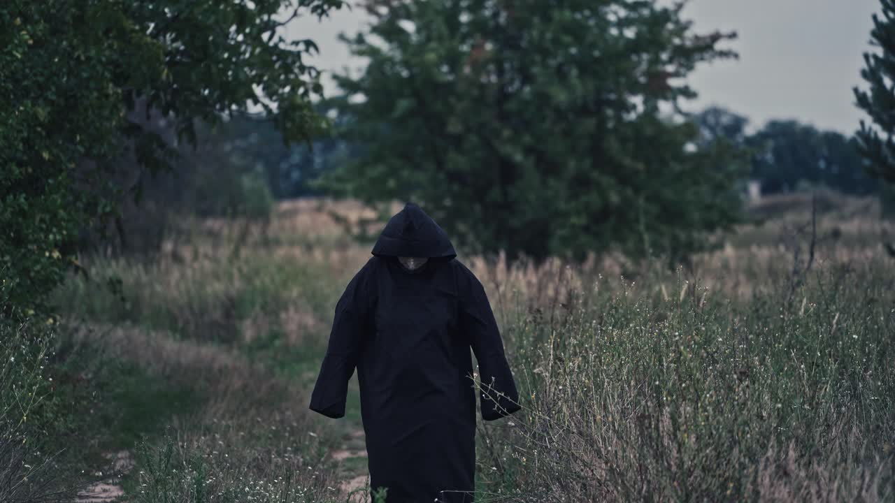 Dark figure in black robe with hood among nature. Scary nightmare walking on the natural background in summer. Mysterious ghost outdoors.