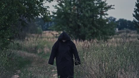 dark figure in black robe with hood among nature. scary nightmare walking on the natural background in summer. mysterious ghost outdoors.