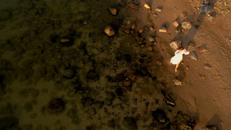 Aerial-Overhead-View-Of-Women-Wearing-Flowing-White-Dress-Walking-Along-Rocky-Beach-Coastline-During-Golden-Hour