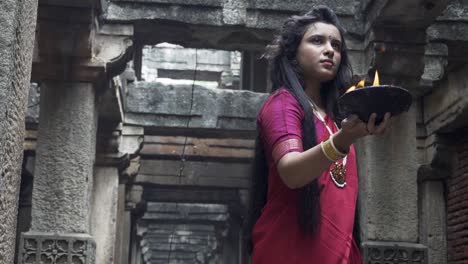 beautiful-Indian-girl-in-old-stepwell-wearing-traditional-Indian-red-saree,-gold-jewellery-and-bangles-holding-fire-plate