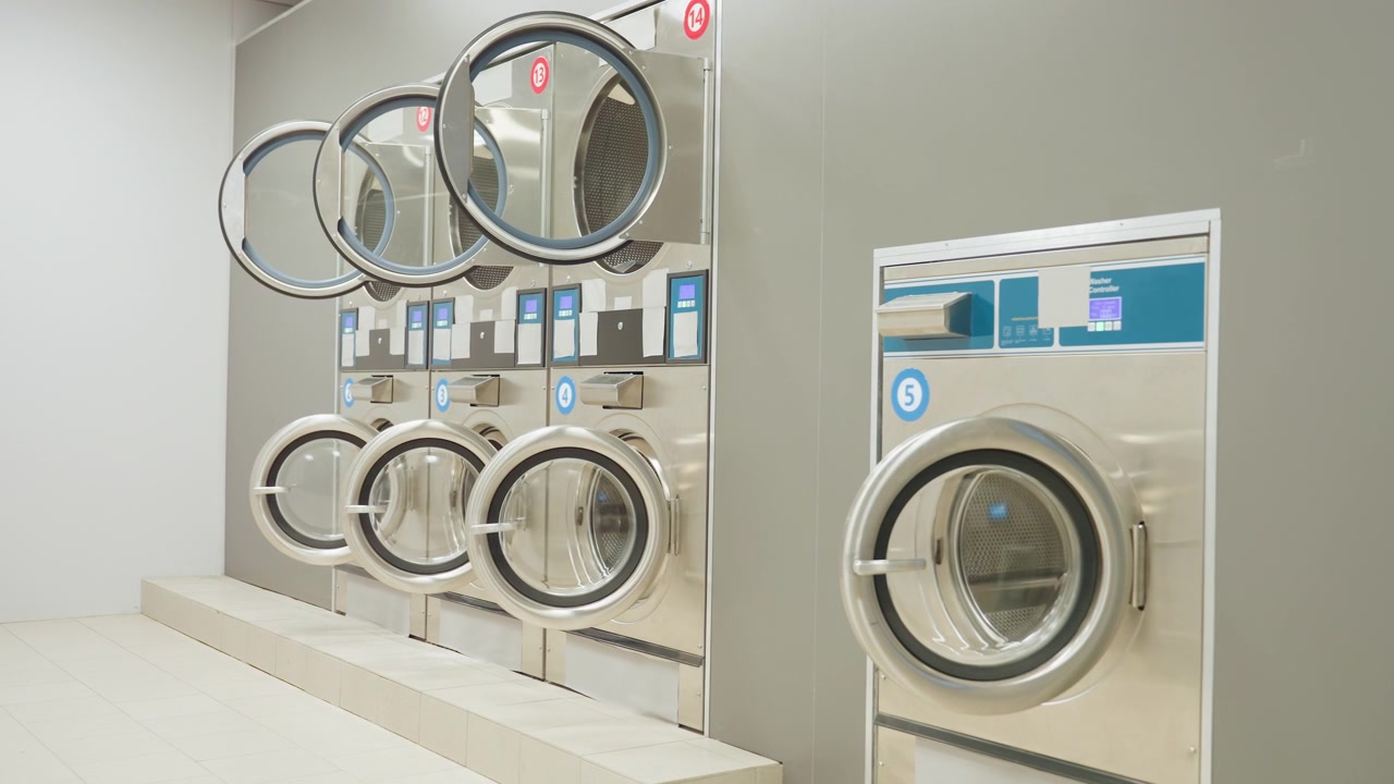 Modern laundry interior with stainless steel washing machines and dryers, doors open, lights switched off, showcasing clean tiled floor, organized baskets, and professional service environment