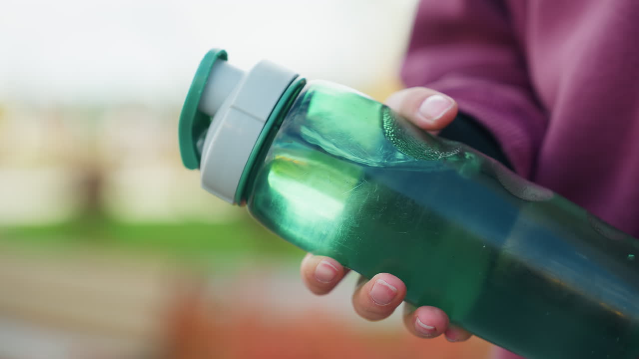Female hand gripping translucent green water bottle with visible swirl and condensation motion against blurred outdoor location conveying dynamic hydration fitness habit energy urban scene