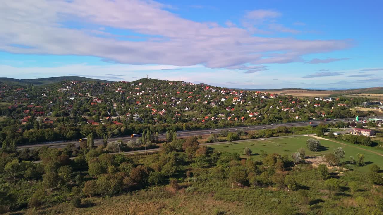 Drone view of cars traveling along the M7 highway in Velence, Hungary, with scenic hillside homes in early autumn sunlight under a partly cloudy sky