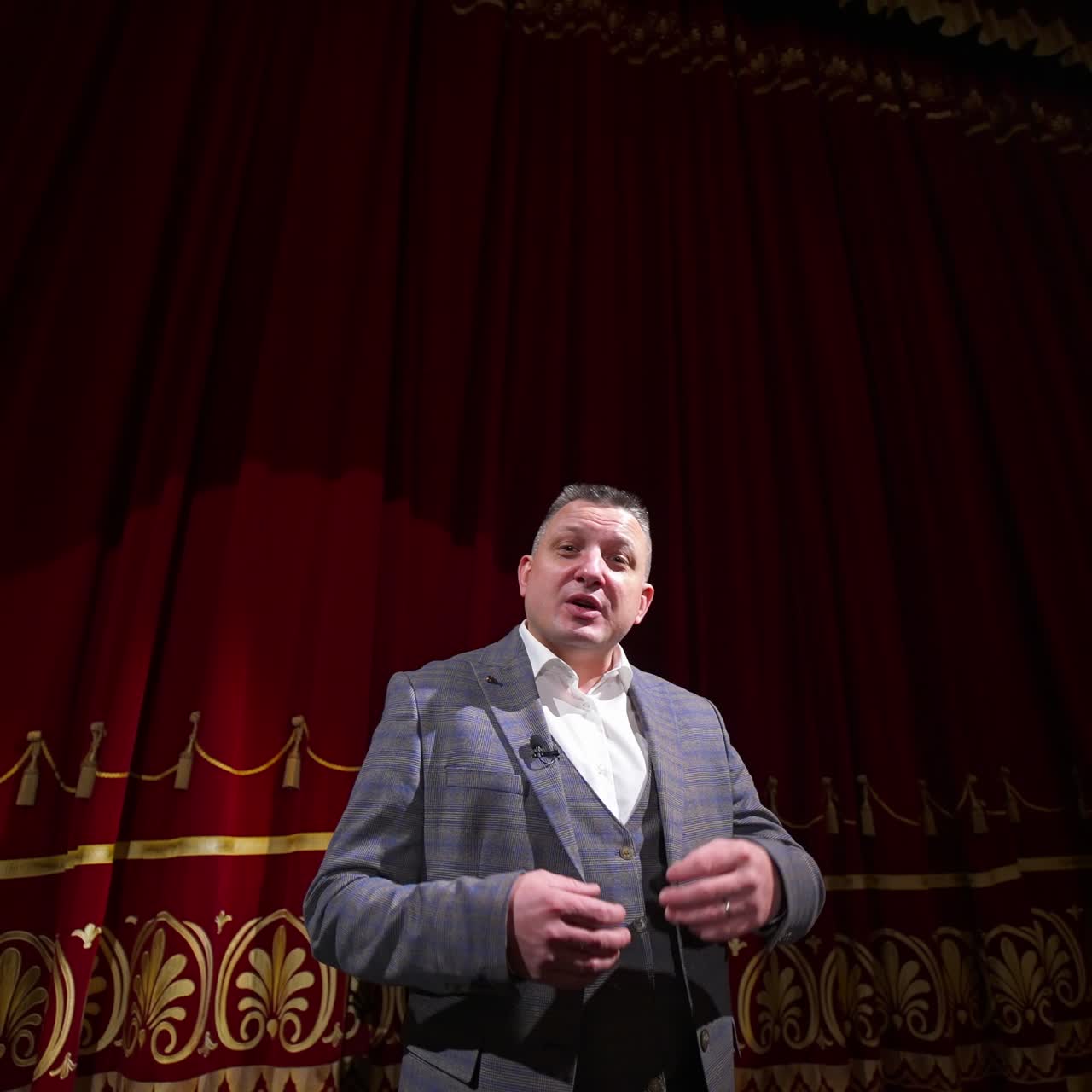 Man in gray costume on stage. Middle-aged actor talking on camera on luxury red curtains background in the theater. Slow motion