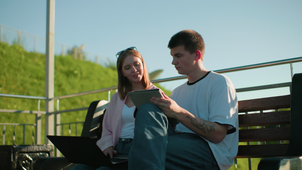 la señora que escribe hace una pausa para mostrar a su amiga sentada cerca de ella mientras él asenta con la cabeza en acuerdo, ambos sentados en un banco al aire libre con barandillas de hierro y colina verde en el fondo en un día soleado