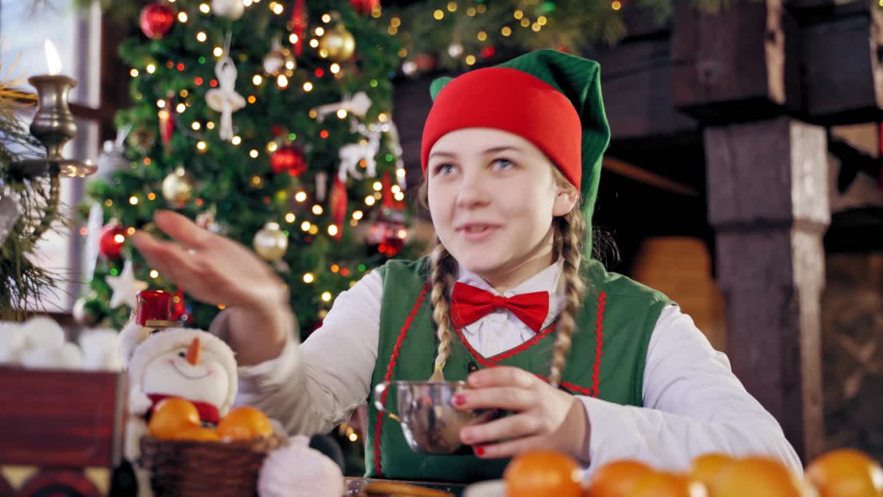 Hungry elf drinking at the table at Christmas. Male elf in green costume with hood eating at festive table on decorated background.