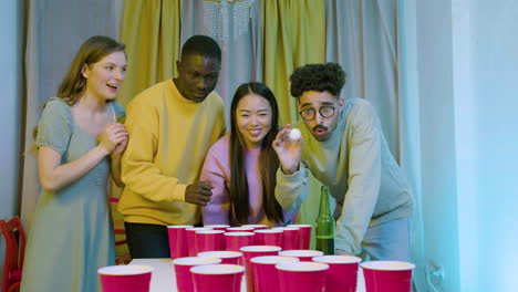 happy multiethnic young friends playing beer pong at home