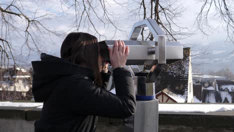 una joven usa binoculares fijos de metal que funcionan con monedas para inspeccionar una pequeña ciudad invernal cubierta de nieve bajo un sol brillante