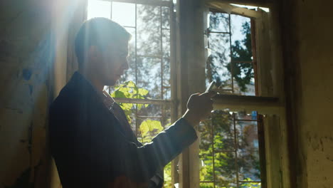 sad young man standing at the window in an abandoned house he looks at the phone's screen concept -