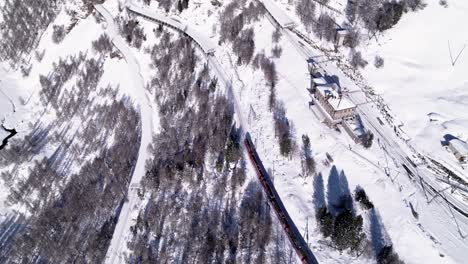 Aerial-view-of-the-Bernina-Express-panorama-train-going-through-a-snow-covered-mountain-winter-landscape-with-forests-on-a-sunny-day-in-Alp-Grum,-Switzerland