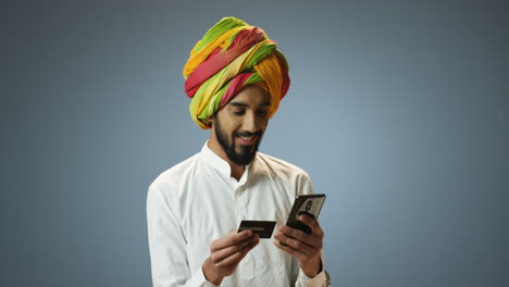 young cheerful indian man in traditional clothes and turban holding credit card and smartphone in hands and shopping online