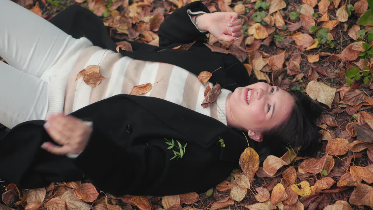 Nature lover lies on forest floor smiling, hands lifting to block falling dry leaves, overhead autumn view with brown foliage and green sprouts framing relaxed playful rest on woodland path