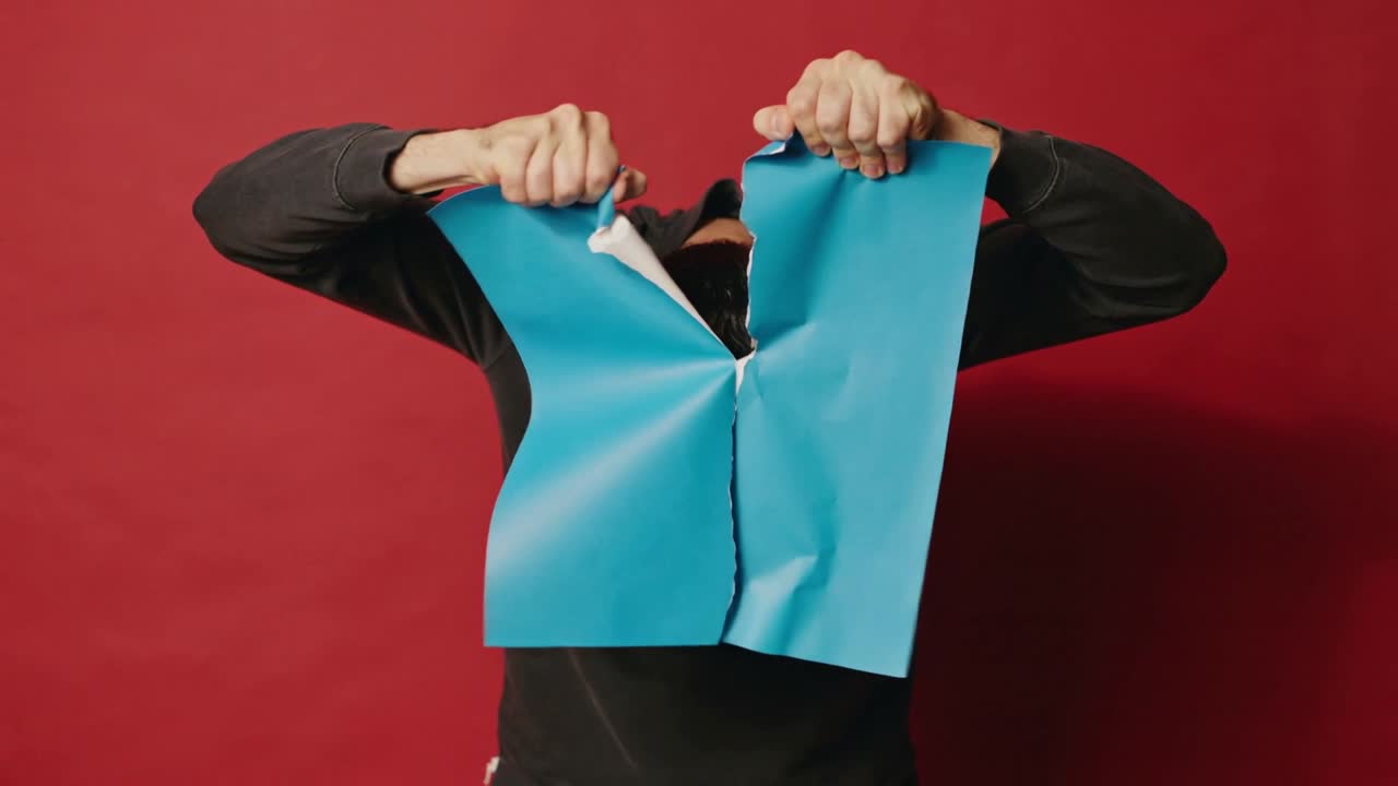 Hombre con suéter oscuro agarrando y rasgando papel azul en un estudio con un telón de fondo rojo, revelando su rostro.
