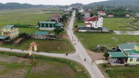 Vista-Aérea-De-Vietnamitas-Conduciendo-Motocicletas-En-La-Carretera-Junto-A-La-Granja-De-Arroz-Y-Las-Casas