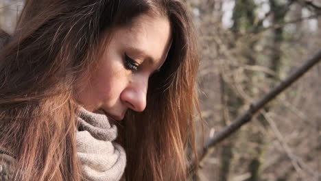 mujer con bufanda de lana mira al suelo en el bosque durante el día soleado en invierno, rayos de sol en su cara - primer plano, toma lateral