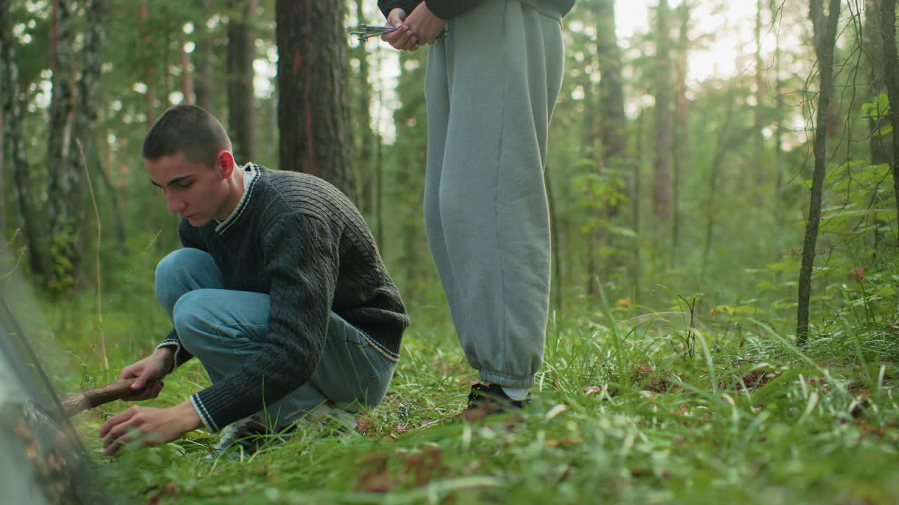 couple squats beside tent with woman handing peg to man holding wood stick preparing to hammer peg into forest ground securing tent among green trees and grass