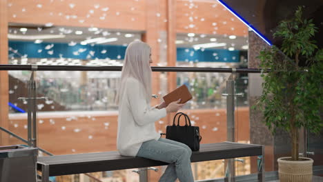 fashionista strides along busy mall promenade, stops beside moving walkway bench, retrieves elegant hardcover from chic tote, delicately flips pages beneath shimmering tree decoration