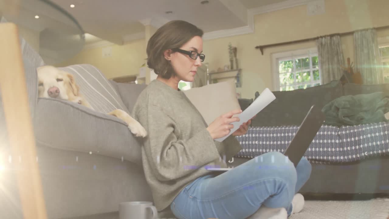 punto de luz contra mujer caucásica con computadora portátil leyendo un documento mientras el perro duerme en el sofá