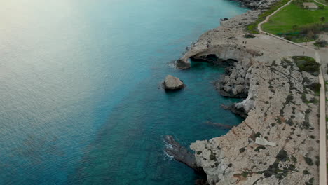 an aerial view of a natural rock archway over the calm blue waters of the mediterranean sea near ayia napa, with a rugged coastline and a grassy area leading up to the arch