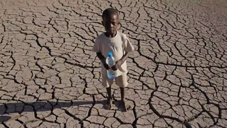 young boy standing on parched, cracked earth, gripping plastic water bottle, symbolizing devastating impact of drought and critical water scarcity in arid landscape