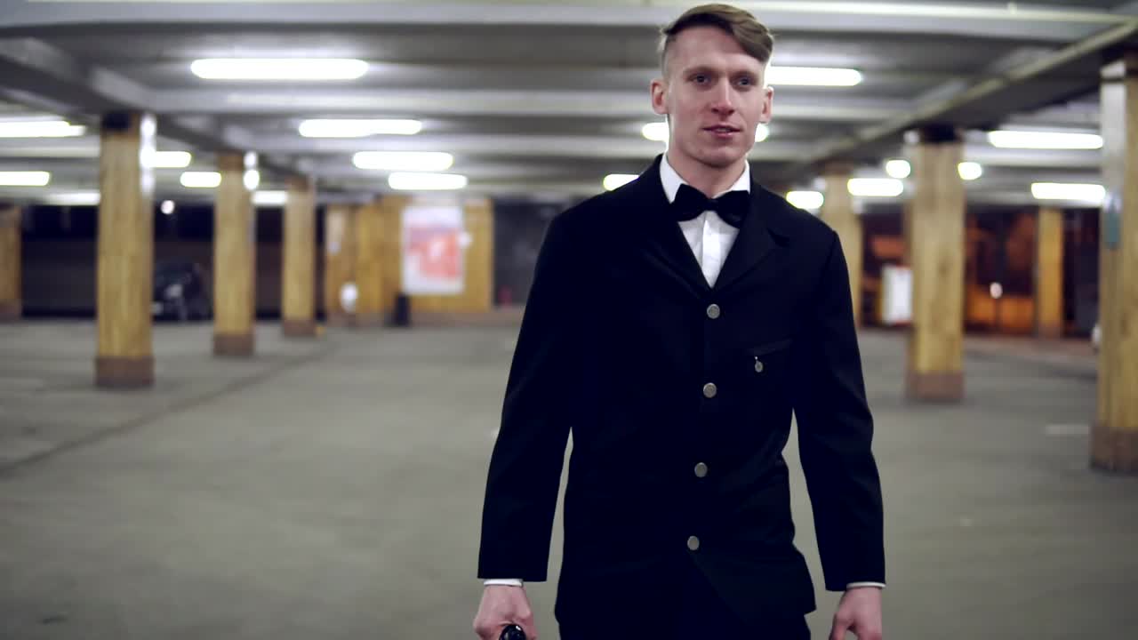 큰 가죽 가방과 함께 주차장에서  ⁇ 고 있는 검은색 양복을 입은 젊은 사업가. 느린 움직임