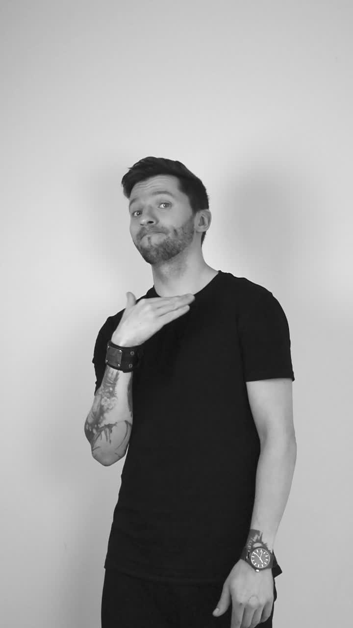 Grayscale footage of white tattooed man with beard and black clothing waving his arm and hand around his neckline or neck indicating someone to stop something and he does not like the thing happening.