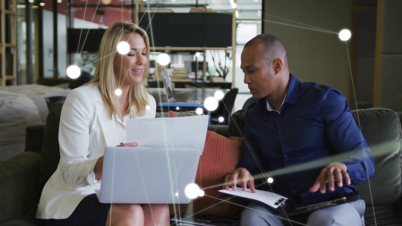 Biz pair exchanging paper and reviewing laptop on sofa, causing network nodes flaring and closing