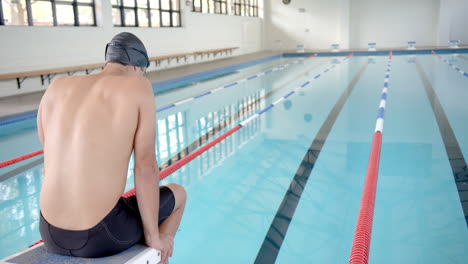 male swimmer in swim cap and goggles sitting on diving block at indoor pool, copy space