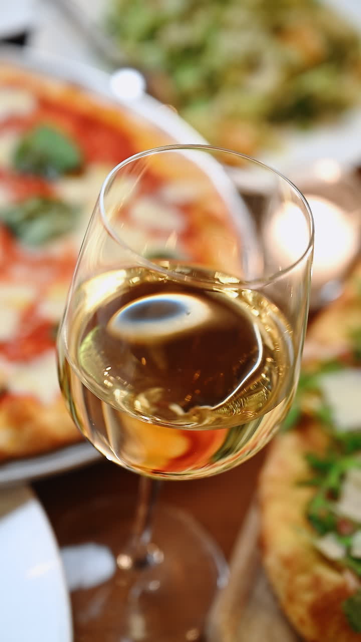 Assaporare una pizza e del vino bianco al tavolo di un ristorante in una giornata di sole. Una persona tiene in mano un bicchiere di vino bianco davanti a una pizza ricoperta di salsa e verdure.