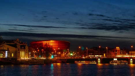 Lapso-De-Tiempo-Del-Tráfico-De-La-Hora-Pico-De-La-Ciudad-Sobre-El-Puente-Del-Tranvía-De-Liepaja,-El-Paisaje-De-La-Ciudad,-Las-Rayas-De-Los-Semáforos,-Los-Tranvías-En-Movimiento-Rápido,-El-Canal-De-La-Ciudad-De-Liepaja-En-Primer-Plano,-Las-Nubes-En-Movimiento,-El-Plano-General