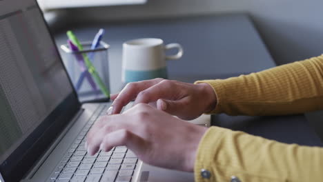 sección media de una mujer biracial sentada en un escritorio usando una computadora portátil y un teléfono inteligente en casa, en cámara lenta