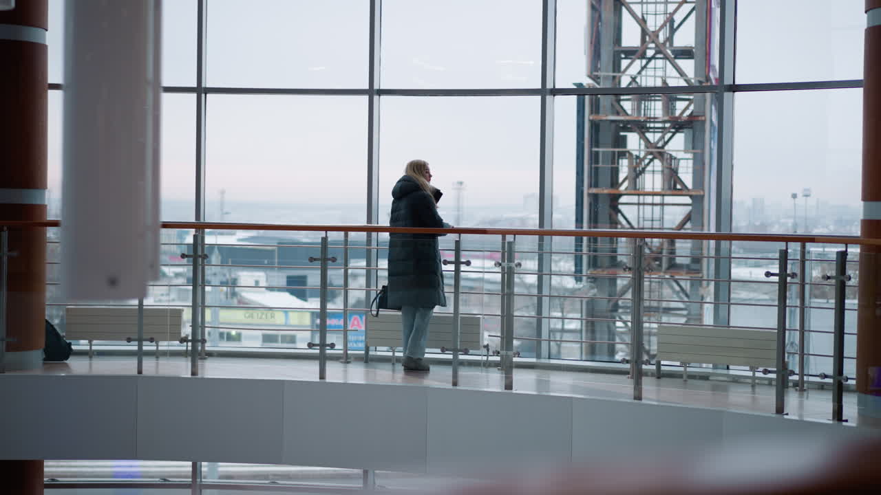 검은색 재을 입은 젊은 여성이 현대 쇼핑몰에서 철 철로에 손을 들고 고 있으며, 뒤에서 흐릿한 도시와 움직이는 자동차가 보이고 있으며, 큰 유리 창문은 도시 풍경을 보여줍니다.