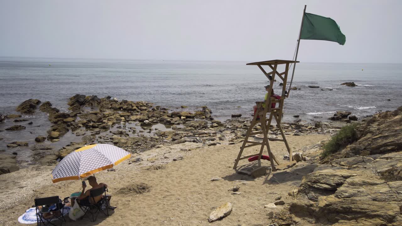 Life guard sits on duty in her post on the beach near Cala de Mijas on the Costa Del Sol in Southern Spain as a holiday makers sits beneath parasol looking at mobile pone.