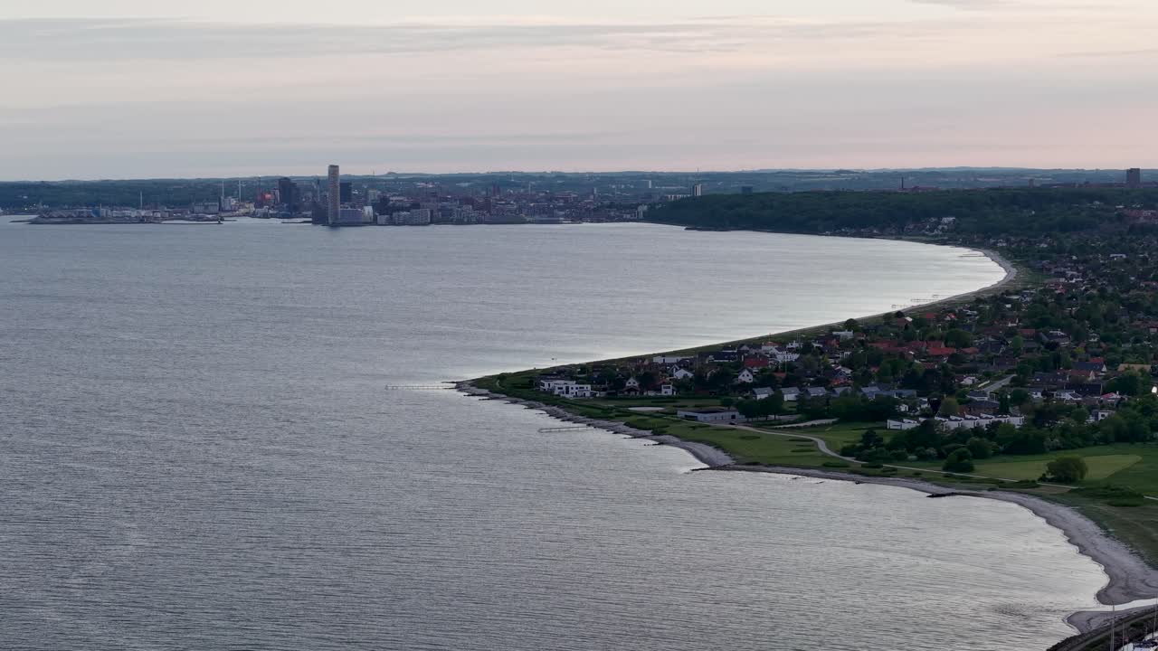 Aerial panorama of a calm bay at sunset with a curving shoreline, adjoining village and distant city skyline under pastel evening skies