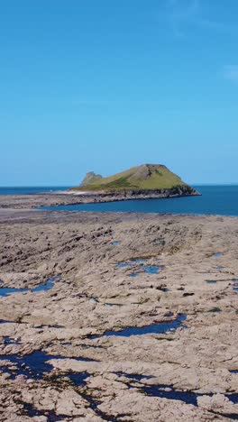 vertical video sideways rising aerial view of worm's head in rhossili bay with rock pool and clear blue sky.