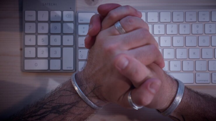 Clasped hands showing extreme nervousness and stress over a keyboard. Close-up shot depicting cybercrime victim or online danger in a moody visual style.