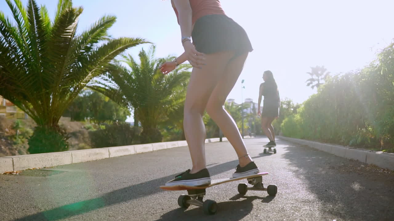 dos chicas en el parque de skate montan a lo largo de la pista a la luz del sol en longboards mirando hacia adelante y riendo en cámara lenta. steadicam vista trasera