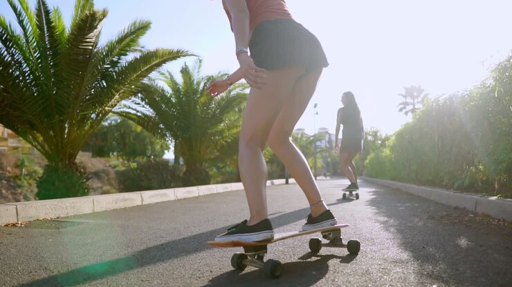 Two girls in skate Park ride along the track in sunlight on longboards looking forward and laughing in slow motion. steadicam back view