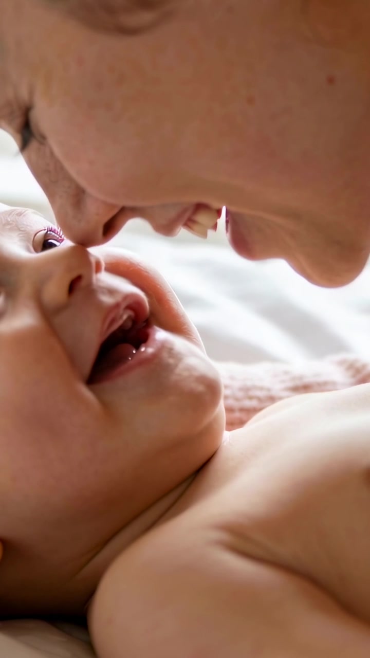 Video verticale: Madre che si china sorridendo e che provoca gorgoglii da un neonato a torso nudo su un letto morbido.