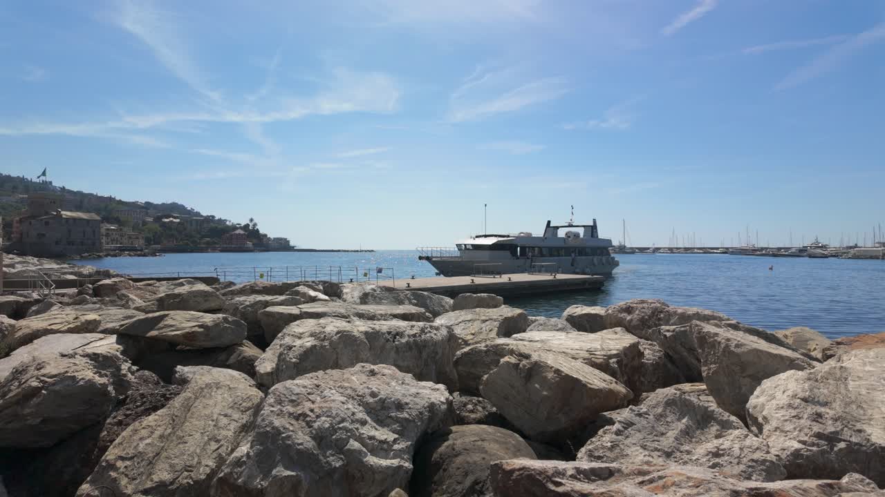Scena di porto tranquilla con grandi rocce in primo piano, uno yacht di lusso ormeggiato al molo, acqua illuminata dal sole, cielo blu e una collina distante. Riprese grandangolari e stabili in pieno giorno.