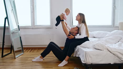 happy parents are together with their infant kid in the bedroom. father raises his baby boy and mom kisses him on the cheek.