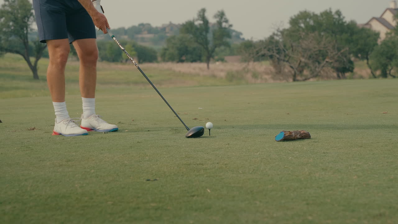 Close-up shot of a ball teed up next to a blue tee marker as a male golfer addresses it with his driver. Framed from the waist down, the scene captures quiet tension and detail before the swing