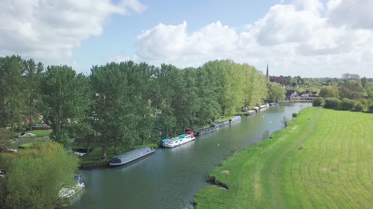 Small Barge And Boats Moored On The River Thames In Abingdon Town Near Oxford city, UK, With Lush Green Trees And Grass. - aerial drone shot