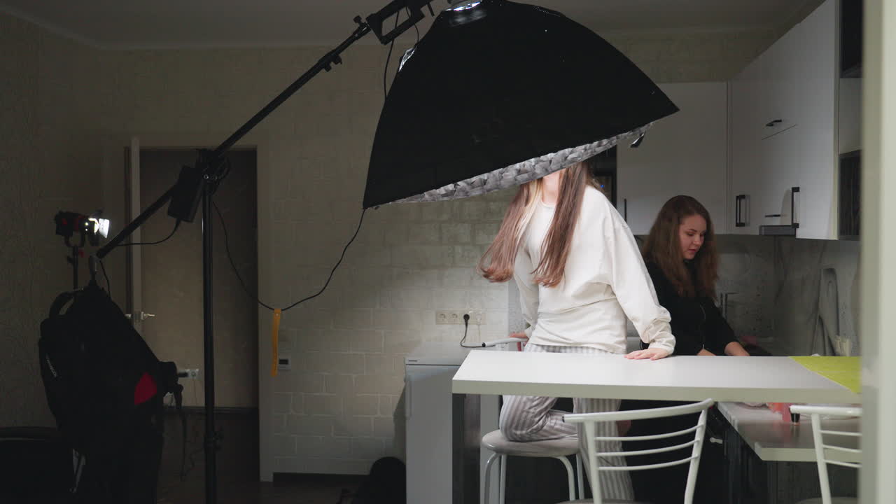 Girlfriends in kitchen space adjusting professional ring light setup, sharing teamwork moment with bright smiles, casual indoor environment showing friendly cooperation in creative activity